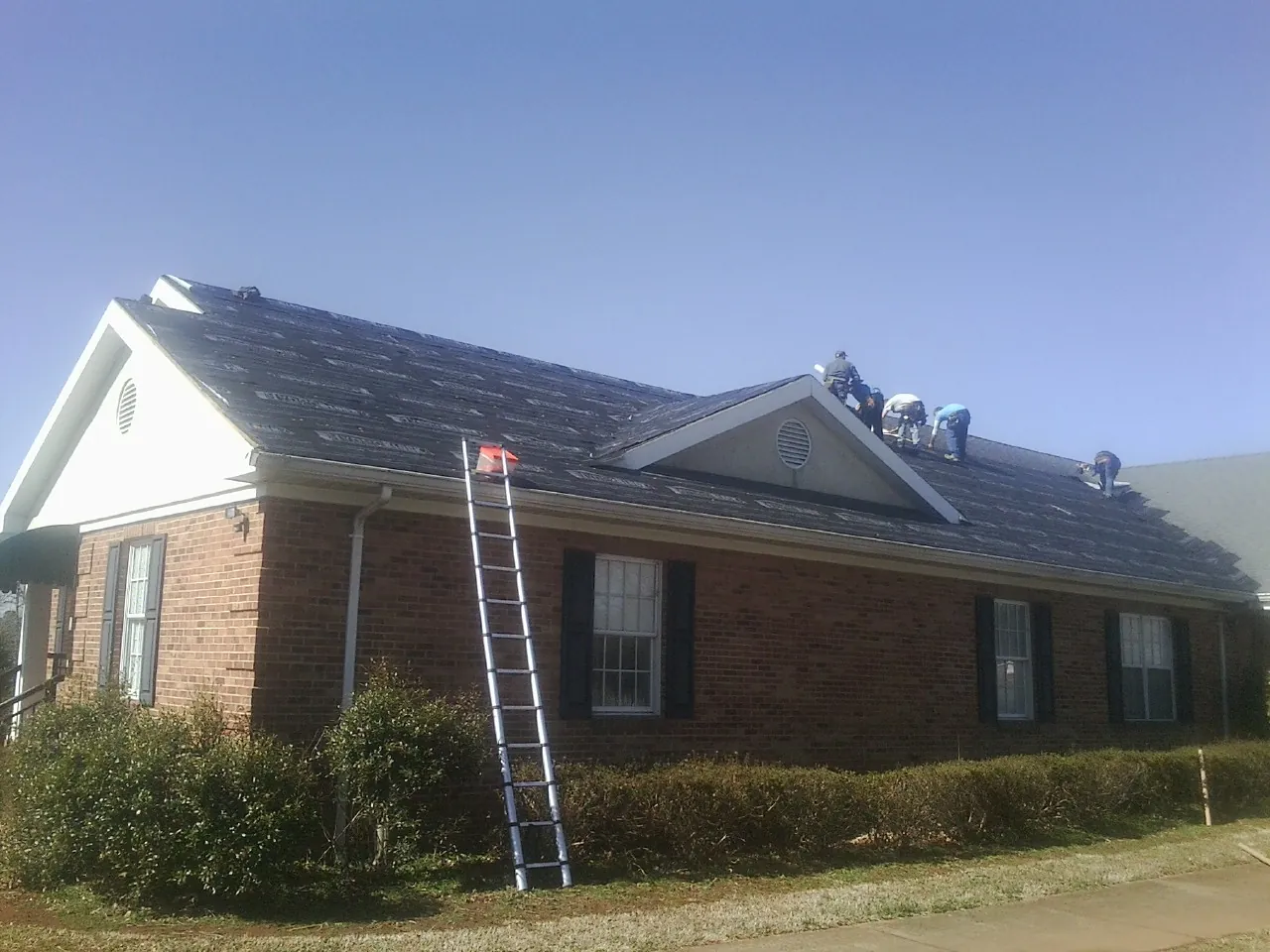 Crew tearing shingles off a brick ranch roof with a ladder set — Emergency Roof Repair work by our crew in Kasson