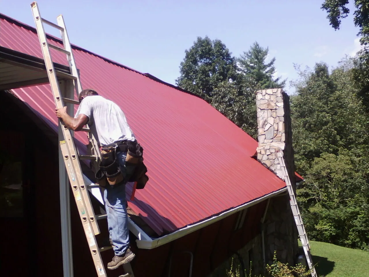 Roofer on a ladder beside a red metal roof and stone chimney — Soffit & Fascia Repair by Roofing Brothers in Kasson